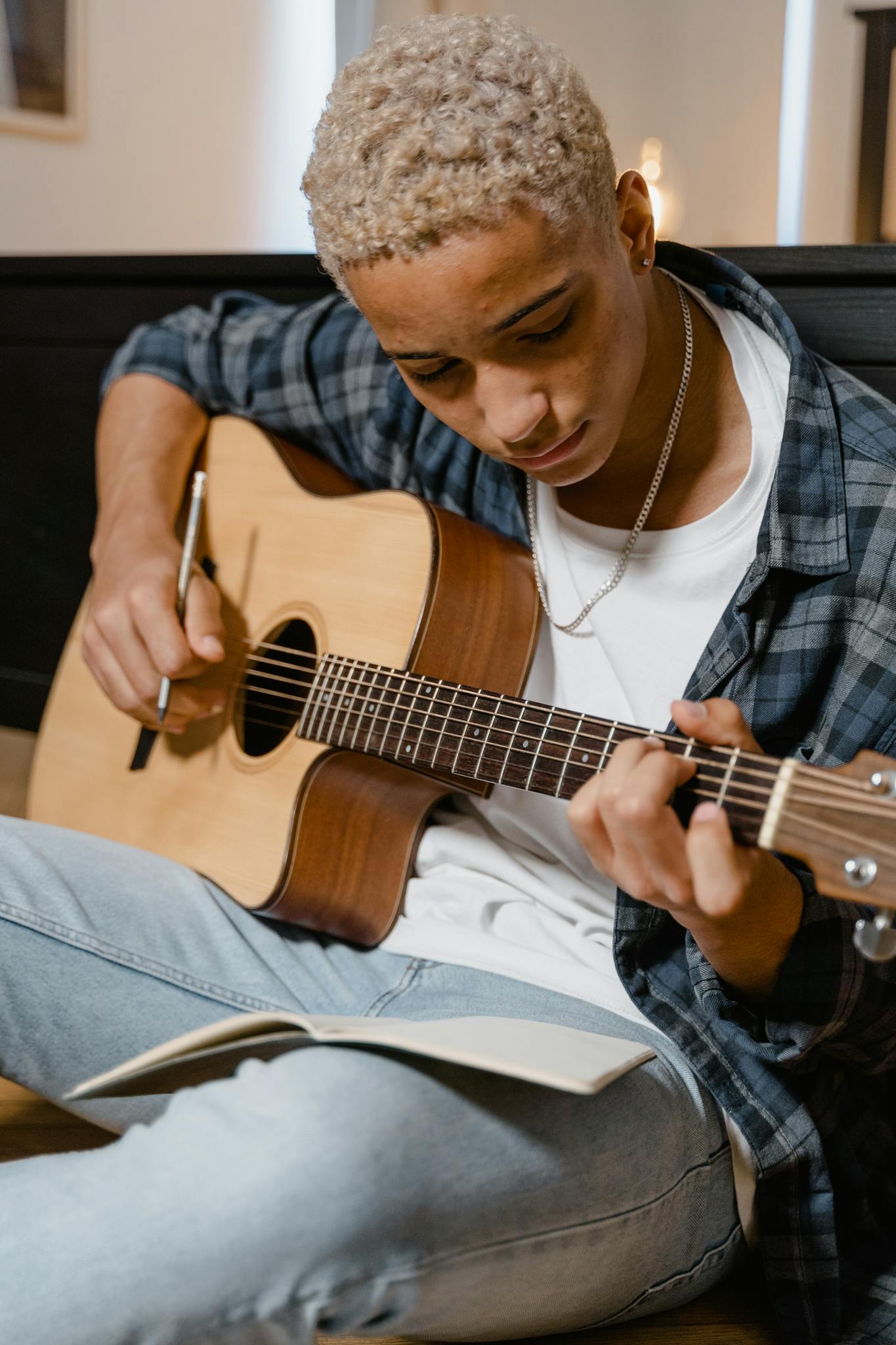 A young man sits in a cozy room playing an acoustic guitar while learning from a notebook.